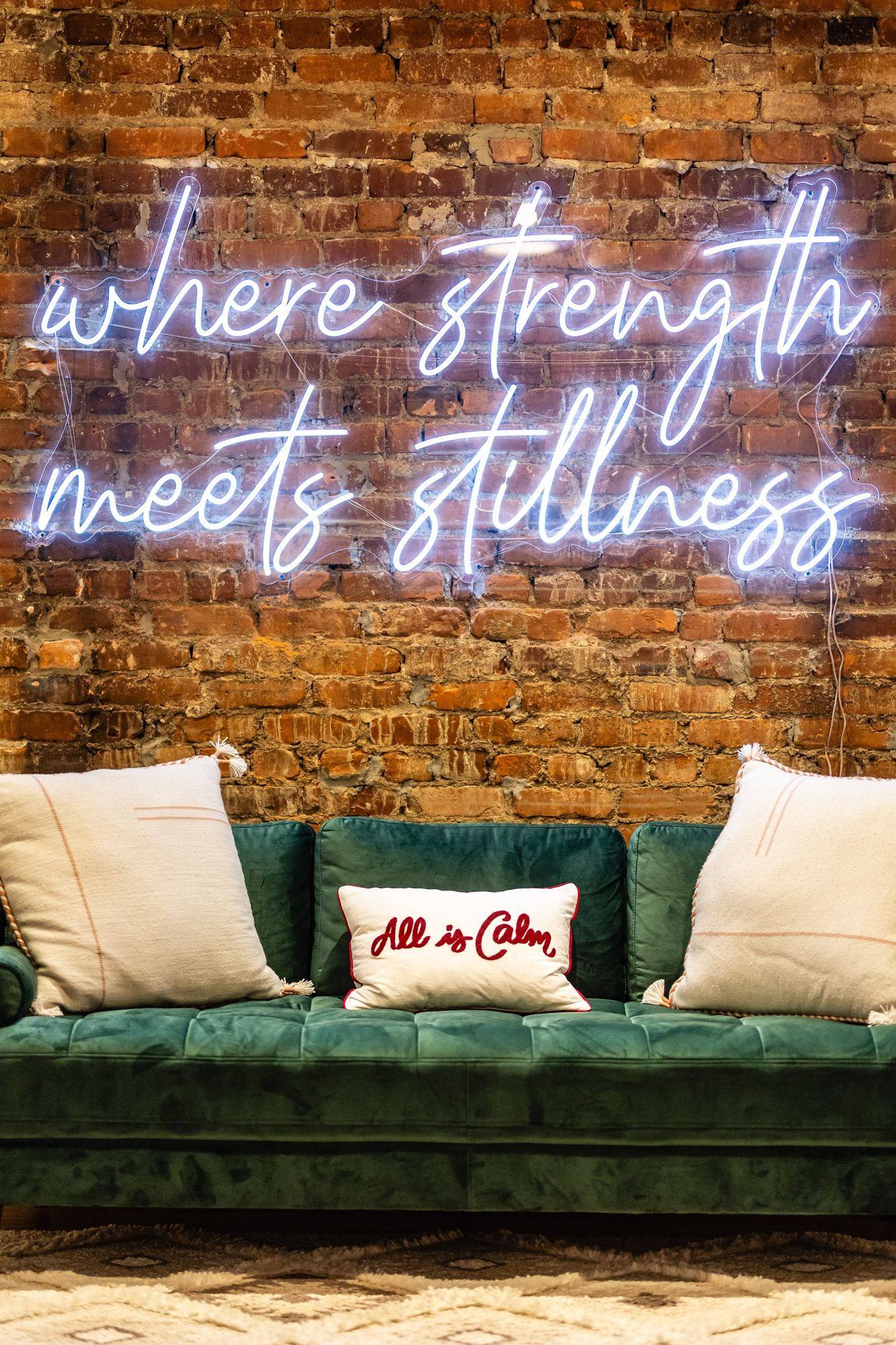Neon sign reading where strength meets stillness above a green velvet sofa